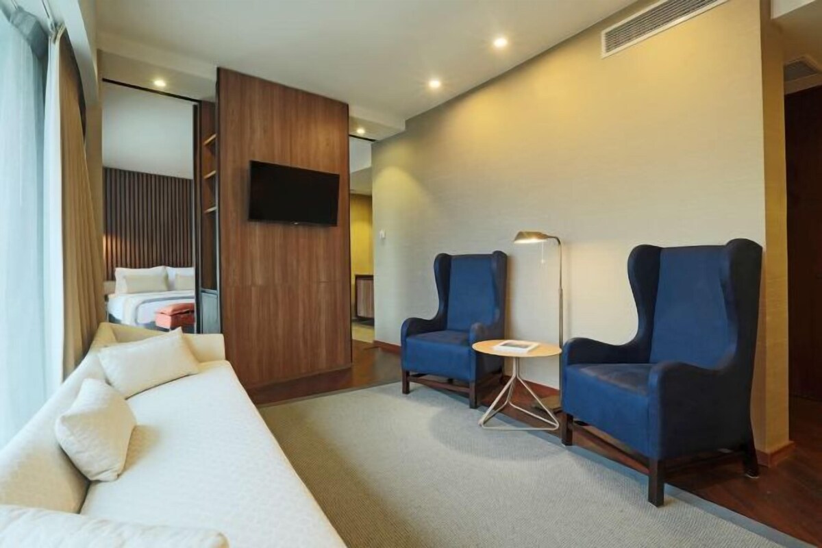 A modern hotel room with a sofa, two armchairs, a small round table, and a flat-screen TV mounted on the wall.