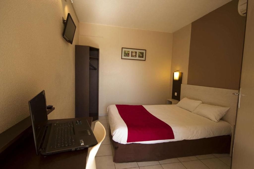 Una camera d'albergo con un letto, una scrivania con un portatile, una TV a parete e un piccolo quadro.