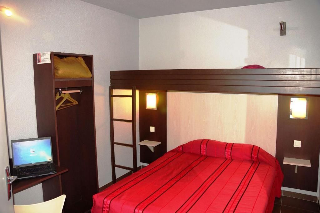 Una cama individual con colcha roja, un ordenador portátil sobre un escritorio y un armario con perchero.