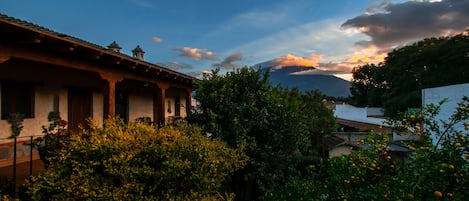 A traditional building with a tiled roof and a bell tower, surrounded by lush greenery and a mountain in the background.