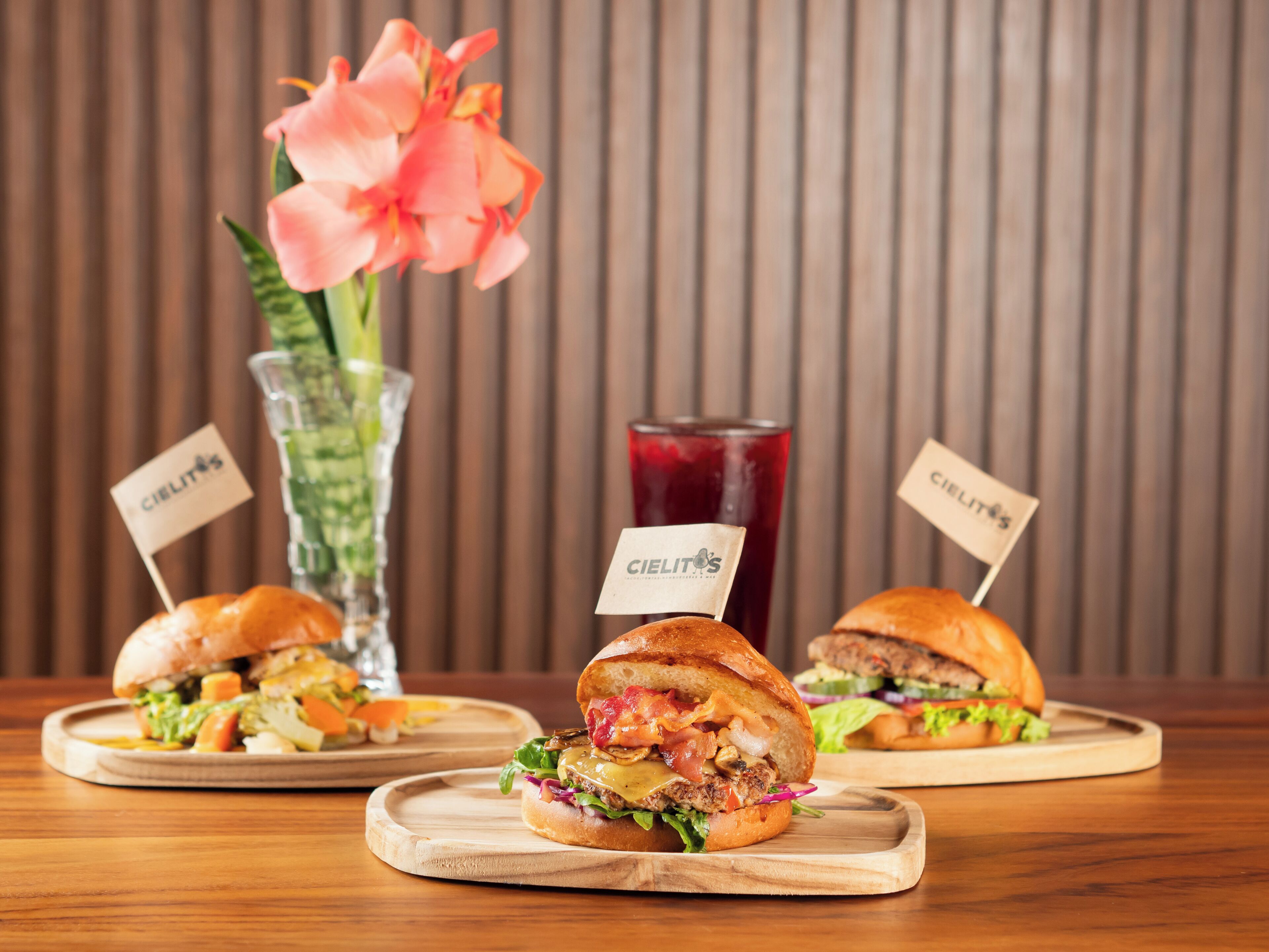 Three sandwiches on wooden boards with 'Cielitos' signs, a glass of red drink, and a pink flower in the background.