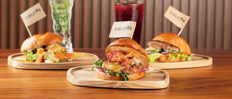 Three sandwiches on wooden boards with 'Cielitos' signs, a glass of red drink, and a pink flower in the background.