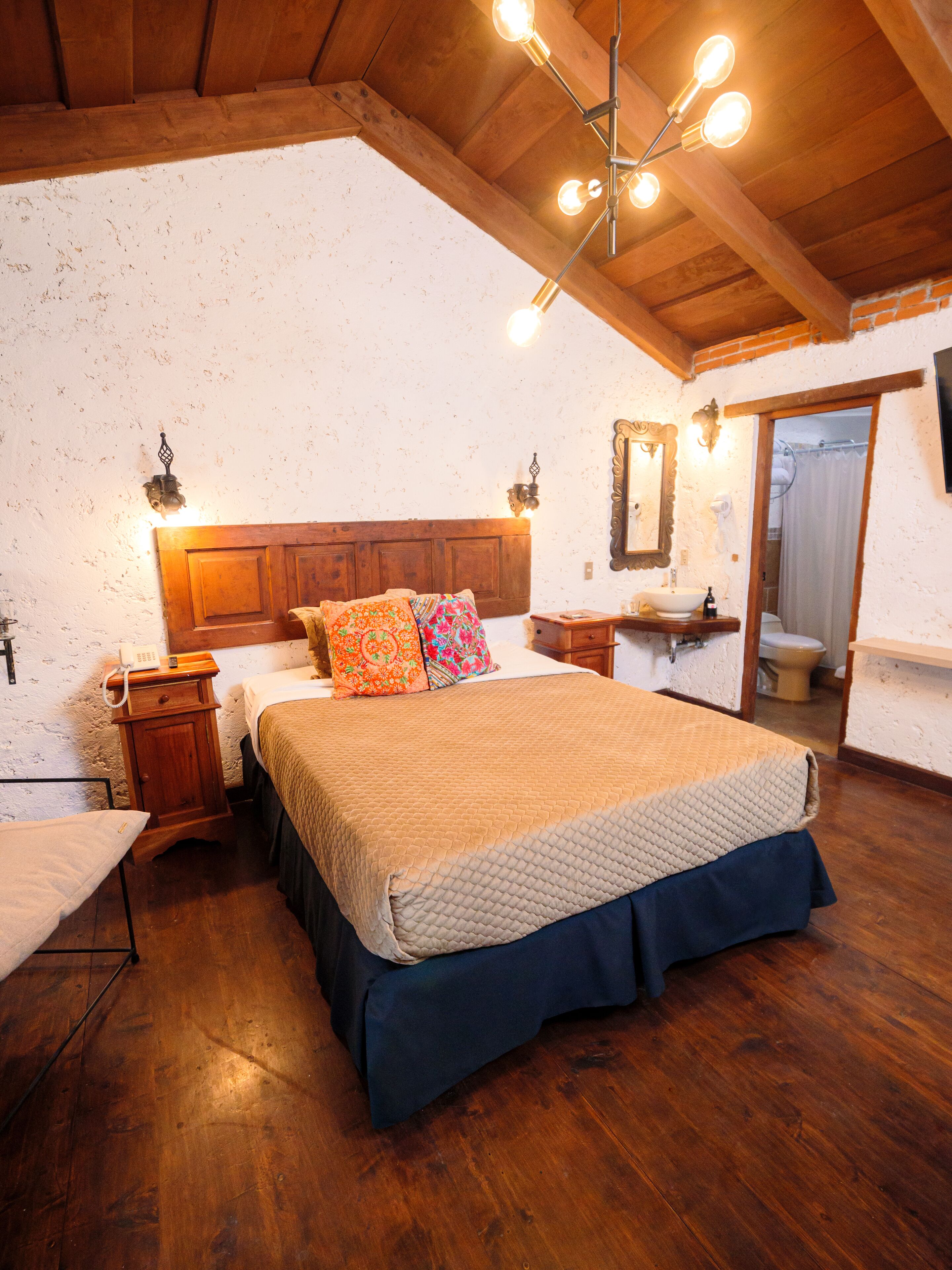 A bedroom with a wooden ceiling, a bed with a quilted cover, a nightstand, a bedside table, and a mirror.