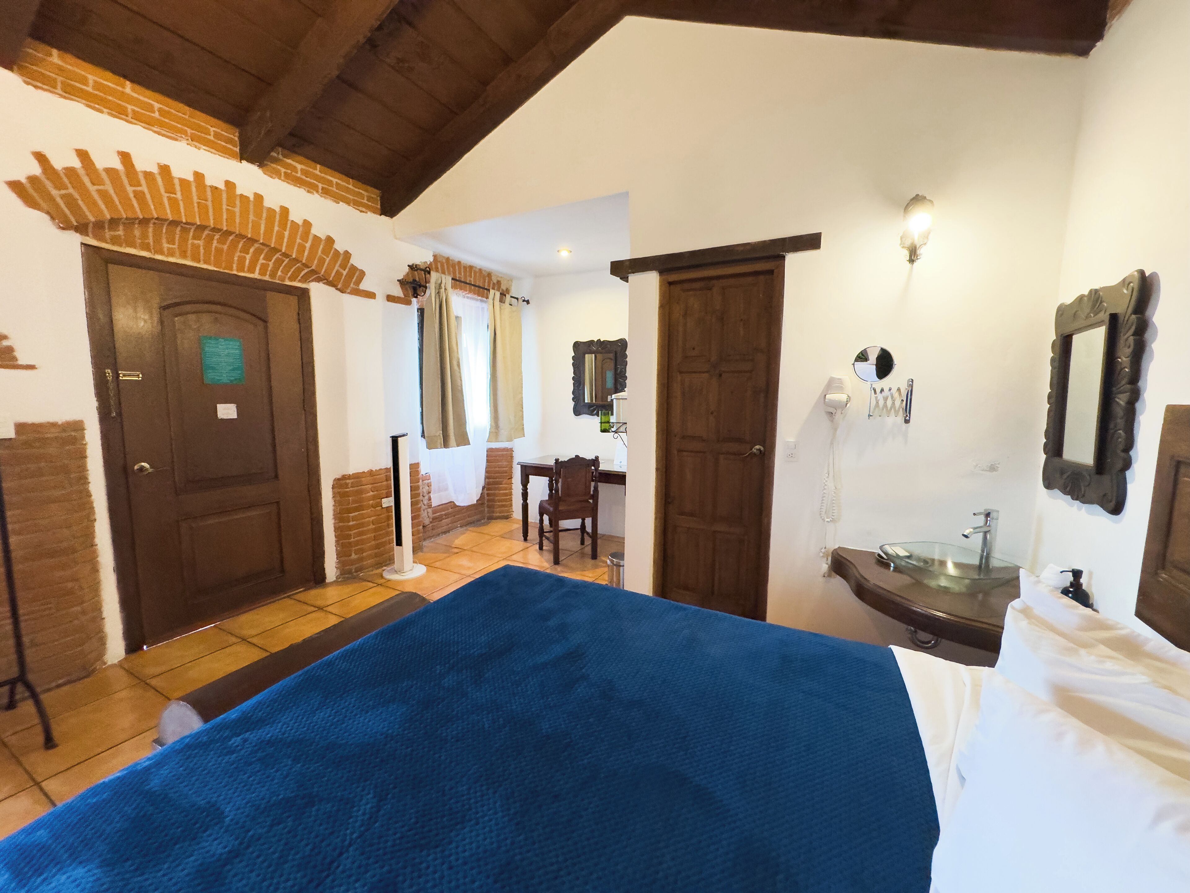 Habitación de hotel con una cama grande, un escritorio, una silla, un baño con lavamanos y espejo, y una puerta que da a otra habitación.