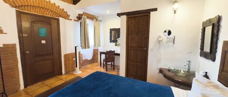 A hotel room with a large bed, a desk, a chair, a bathroom with a sink and mirror, and a door leading to another room.