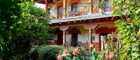 Un edificio de dos pisos con un balcón, rodeado de vegetación exuberante y plantas con flores.