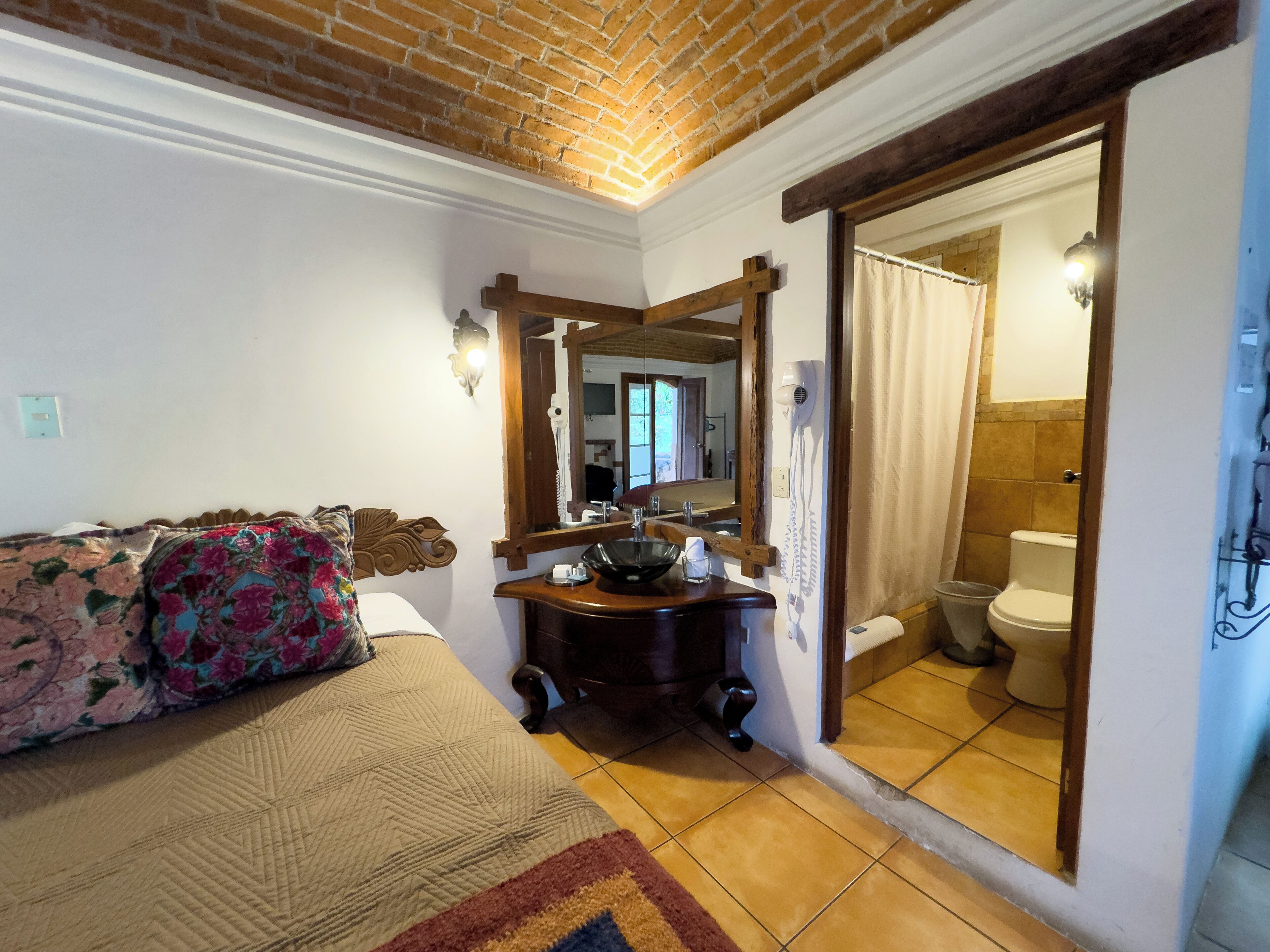 Un dormitorio con cama, cómoda de madera, espejo y baño visible a través de una puerta abierta.