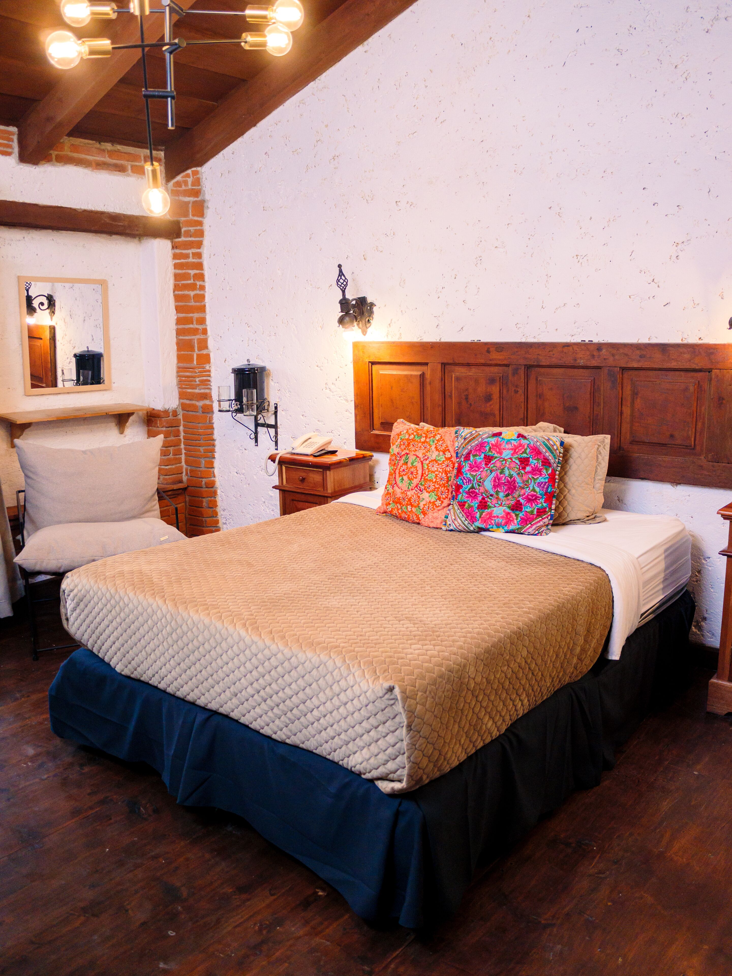 A bedroom with a wooden headboard, a quilted bedspread, a chair, a nightstand, and a wall-mounted lamp.