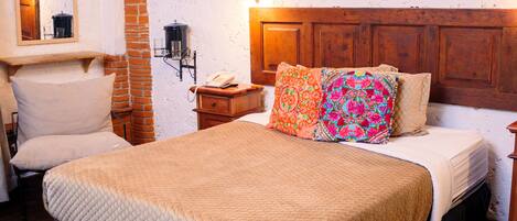 A bedroom with a wooden headboard, a quilted bedspread, a chair, a nightstand, and a wall-mounted lamp.