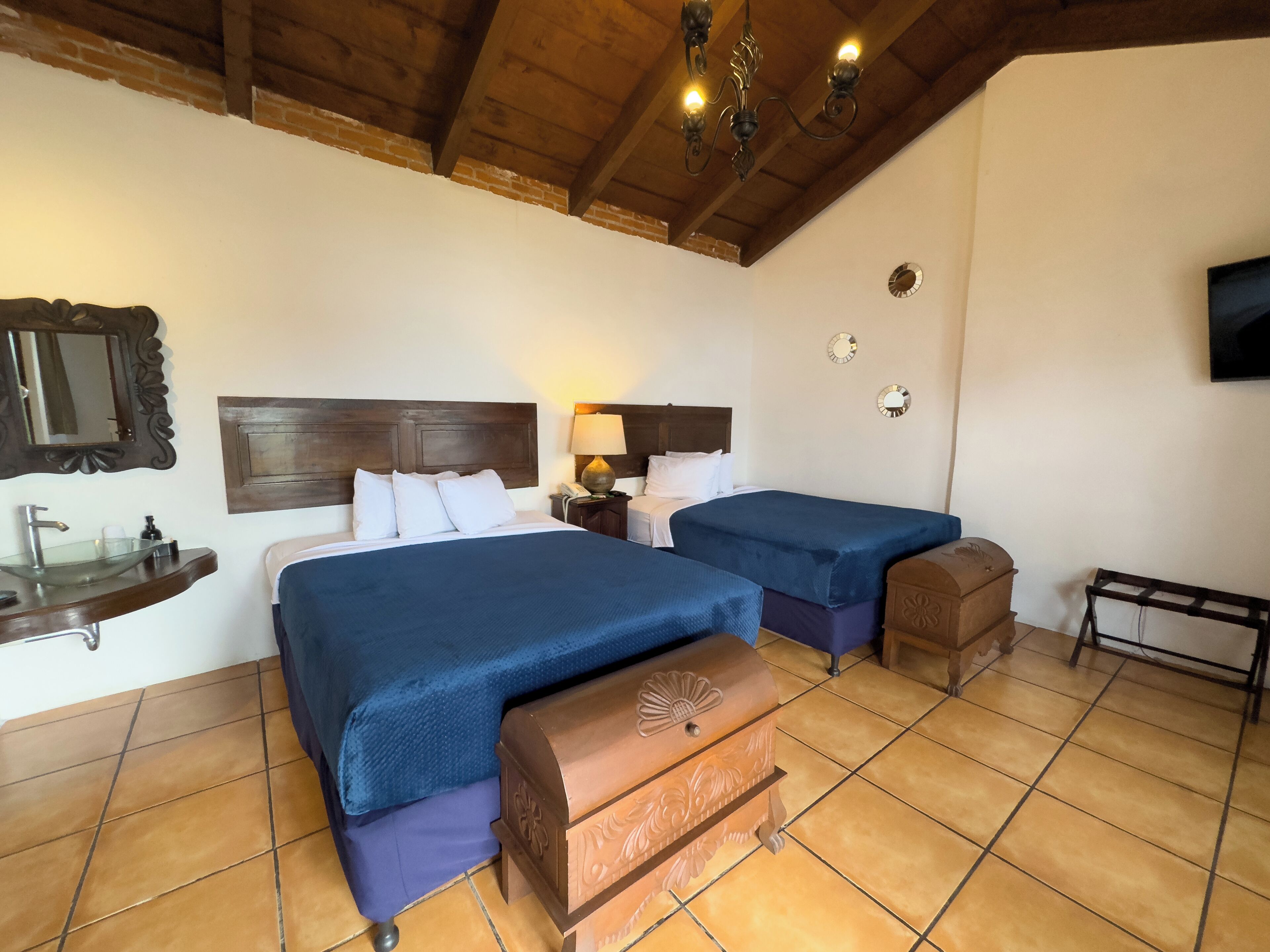 Habitación de hotel con dos camas, cabecero de madera, mesita de noche con lámpara, pequeño cofre de madera, espejo y televisor en la pared.