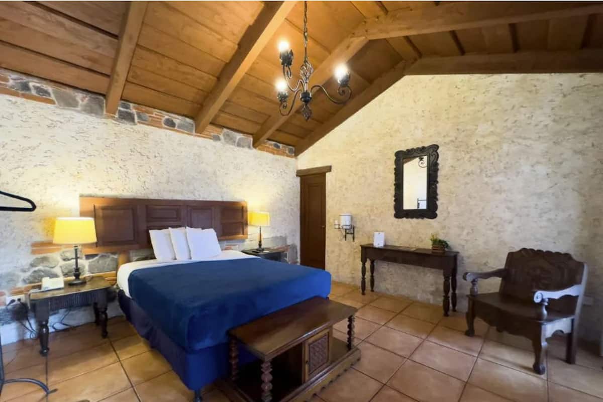 Ein Schlafzimmer mit Holzboden, Steinwänden, einem Bett mit blauer Bettdecke, zwei Nachttischlampen, einer hölzernen Bank, einem Stuhl und einem Spiegel an der Wand.