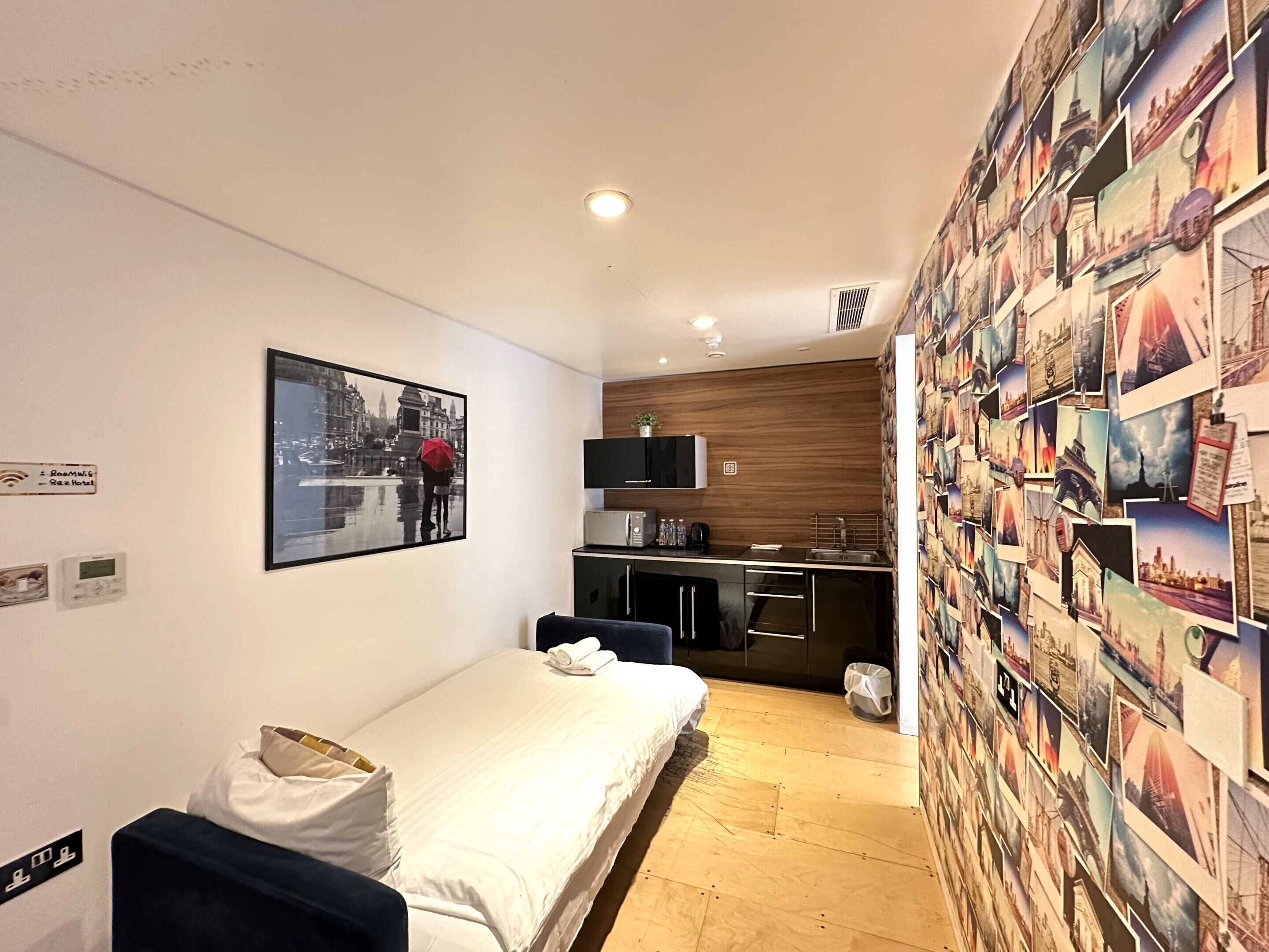 A compact hotel room with a bed, a small kitchenette, a wall adorned with travel photos, and a cityscape artwork.