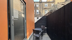 A narrow outdoor space with a sliding glass door, a small table with two chairs, and a black fence. - Room Home Stay (London)