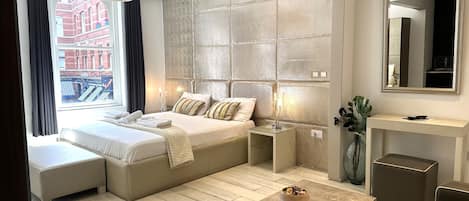 A modern hotel room with a large bed, a seating area, a bedside table with a lamp, a mirror, and a view of the city.