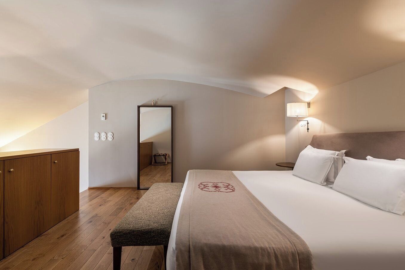 Ein Schlafzimmer mit einem Bett, einem Holzschrank, einer Bank, einem Spiegel und einem Nachttisch mit Lampe.