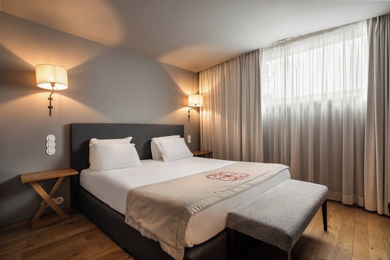 A hotel room with a large bed, two bedside lamps, a bench, and a view of the outdoors through sheer curtains.