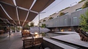 Un espace de restauration en plein air avec des tables et des chaises en bois, un élément décoratif aquatique et un bâtiment moderne aux lignes verticales en arrière-plan.