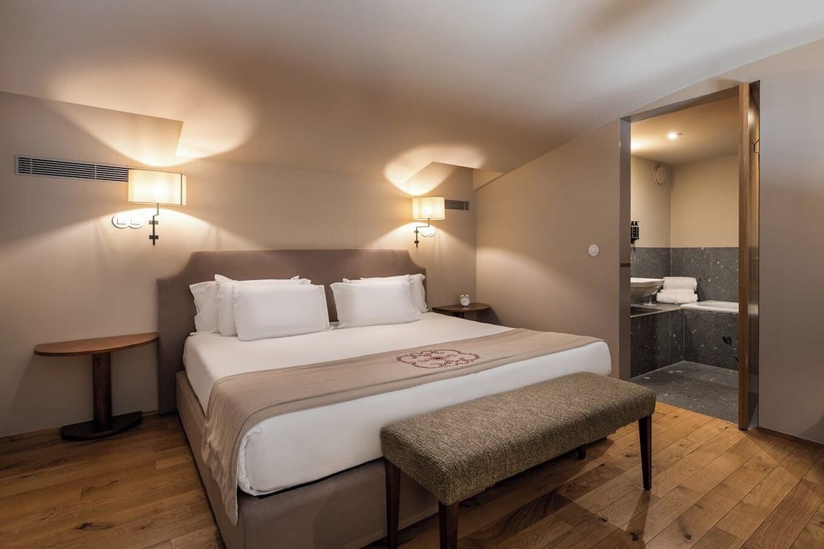 Een hotelkamer met een bed, nachtkastjes, een bankje en een badkamer zichtbaar door een geopende deur.
