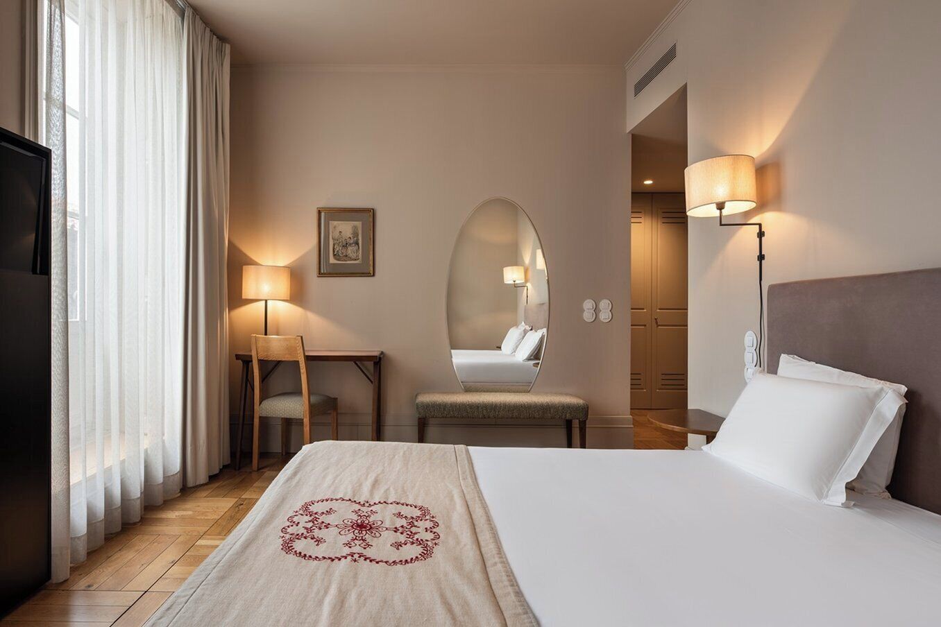 A hotel room with a bed, a desk, a chair, a mirror, and a lamp.