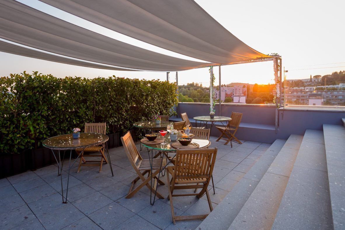 An outdoor patio with a glass table and wooden chairs, surrounded by greenery and overlooking a cityscape.