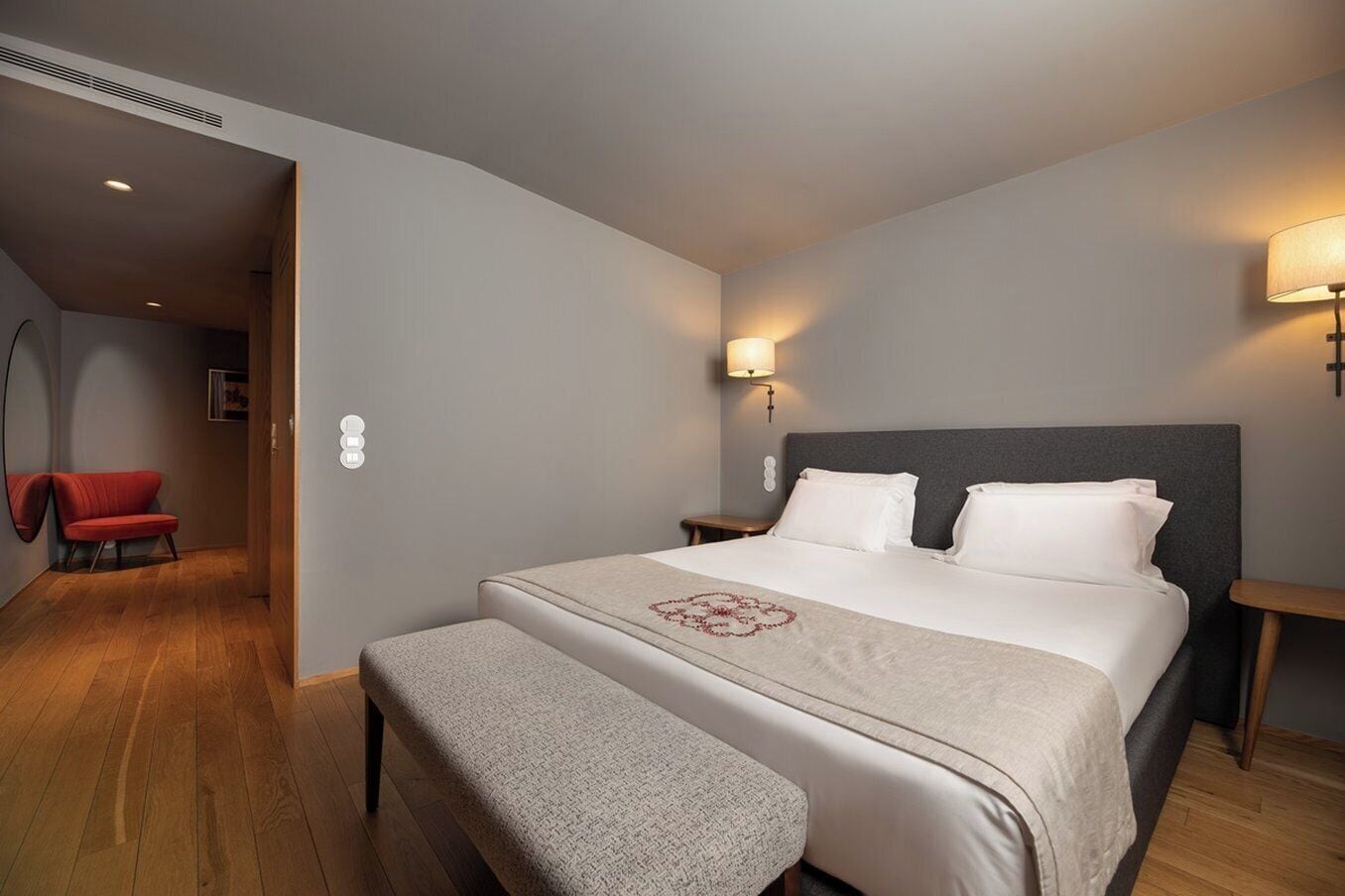Une chambre d’hôtel avec un grand lit, une banquette, un fauteuil rouge et deux lampes fixées au mur.