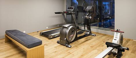 A gym with a treadmill, elliptical, and rowing machine, a bench, and a large window with a view of trees.