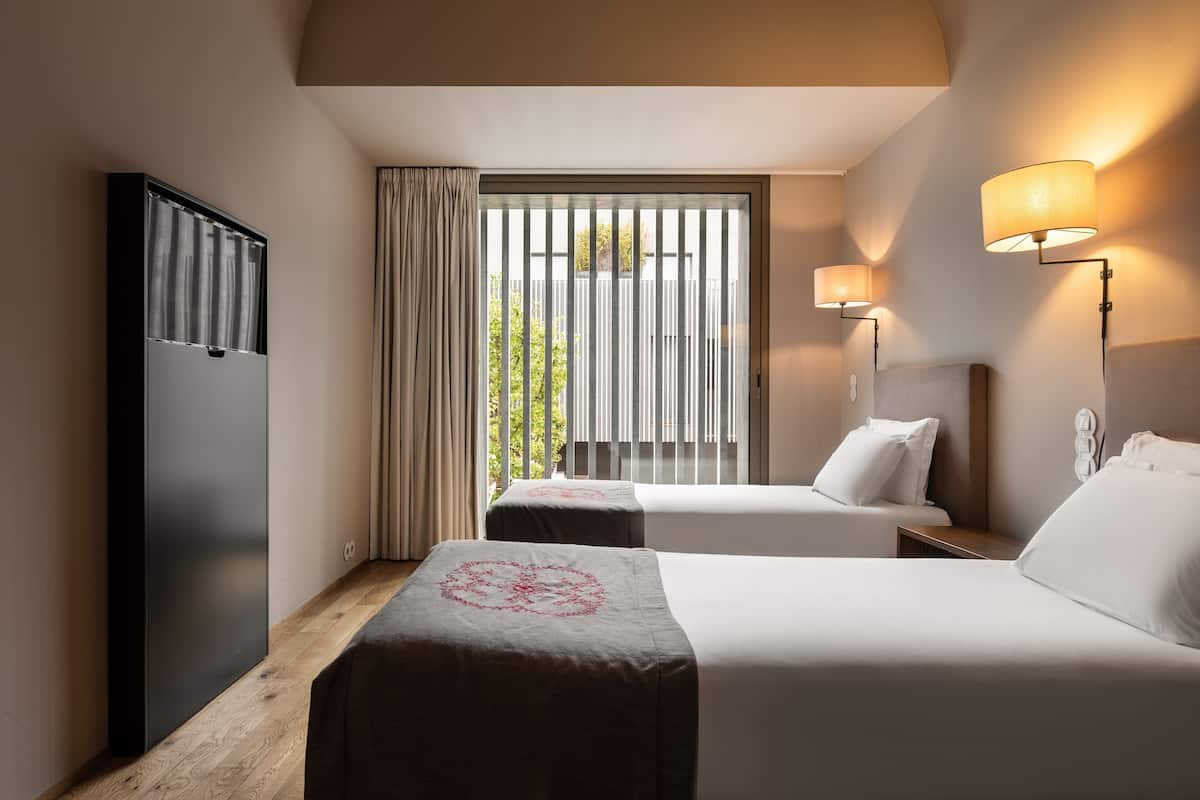 Een hotelkamer met twee bedden, een kledingkast, een raam met zonwering en wandlampen.