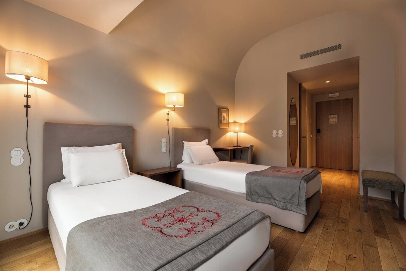 Une chambre d’hôtel avec deux lits, un bureau et une chaise. On y trouve des lampes fixées au mur et une porte portant un numéro.