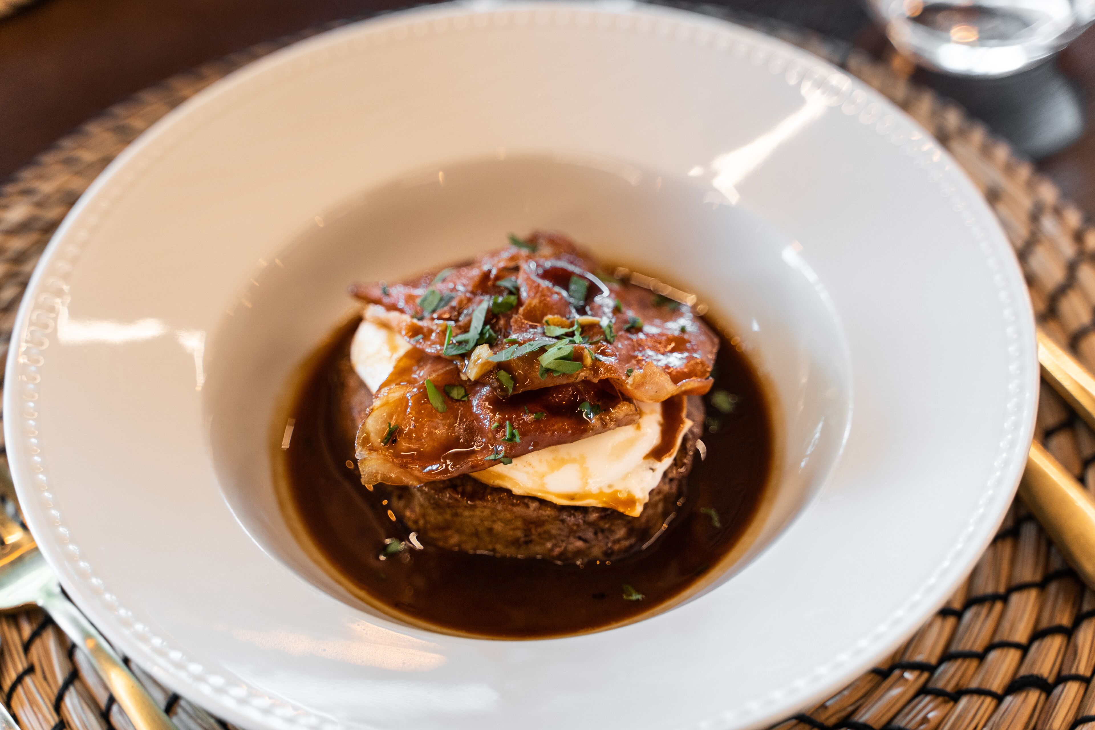 Een witte schaal met een donkerbruin gerecht, mogelijk een steak, getopt met een romige saus en versierd met kruiden.