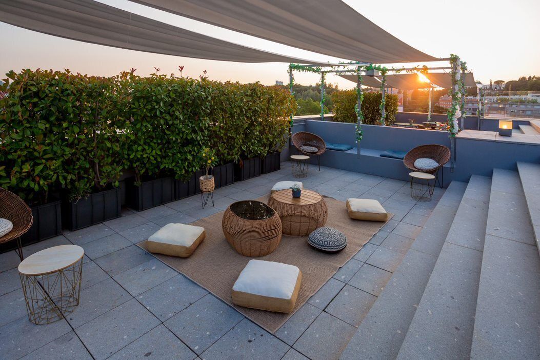 A rooftop patio with outdoor seating, including wicker chairs and ottomans, surrounded by greenery and a pergola.