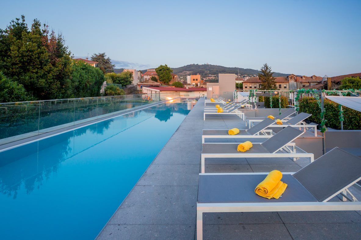 A rooftop pool with clear blue water, surrounded by lounge chairs with yellow towels, set against a backdrop of buildings and trees.