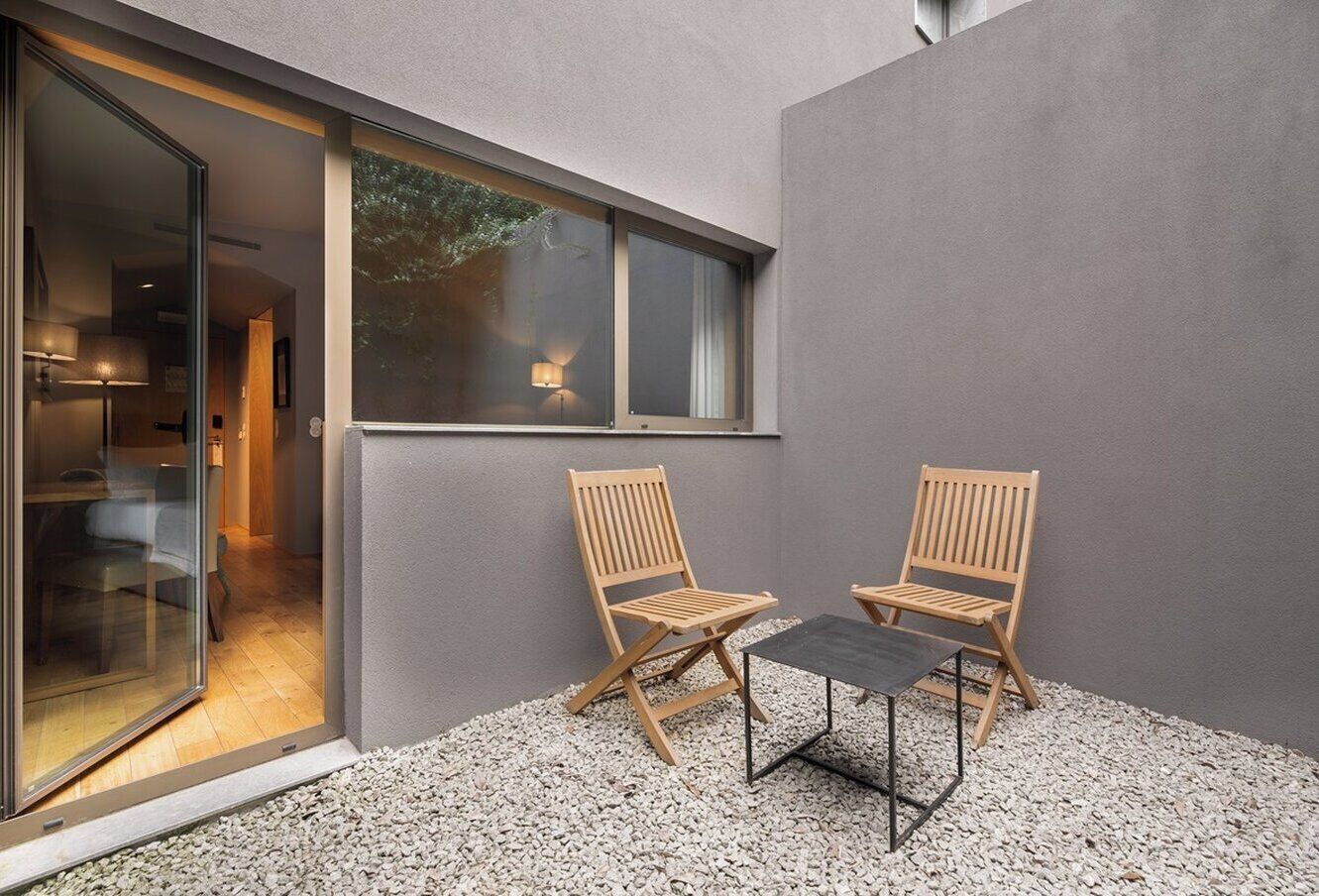 Un espace extérieur moderne aménagé avec deux chaises en bois et une table, entouré de gravier, à côté d’un bâtiment doté d’une porte en verre donnant accès à un espace intérieur.