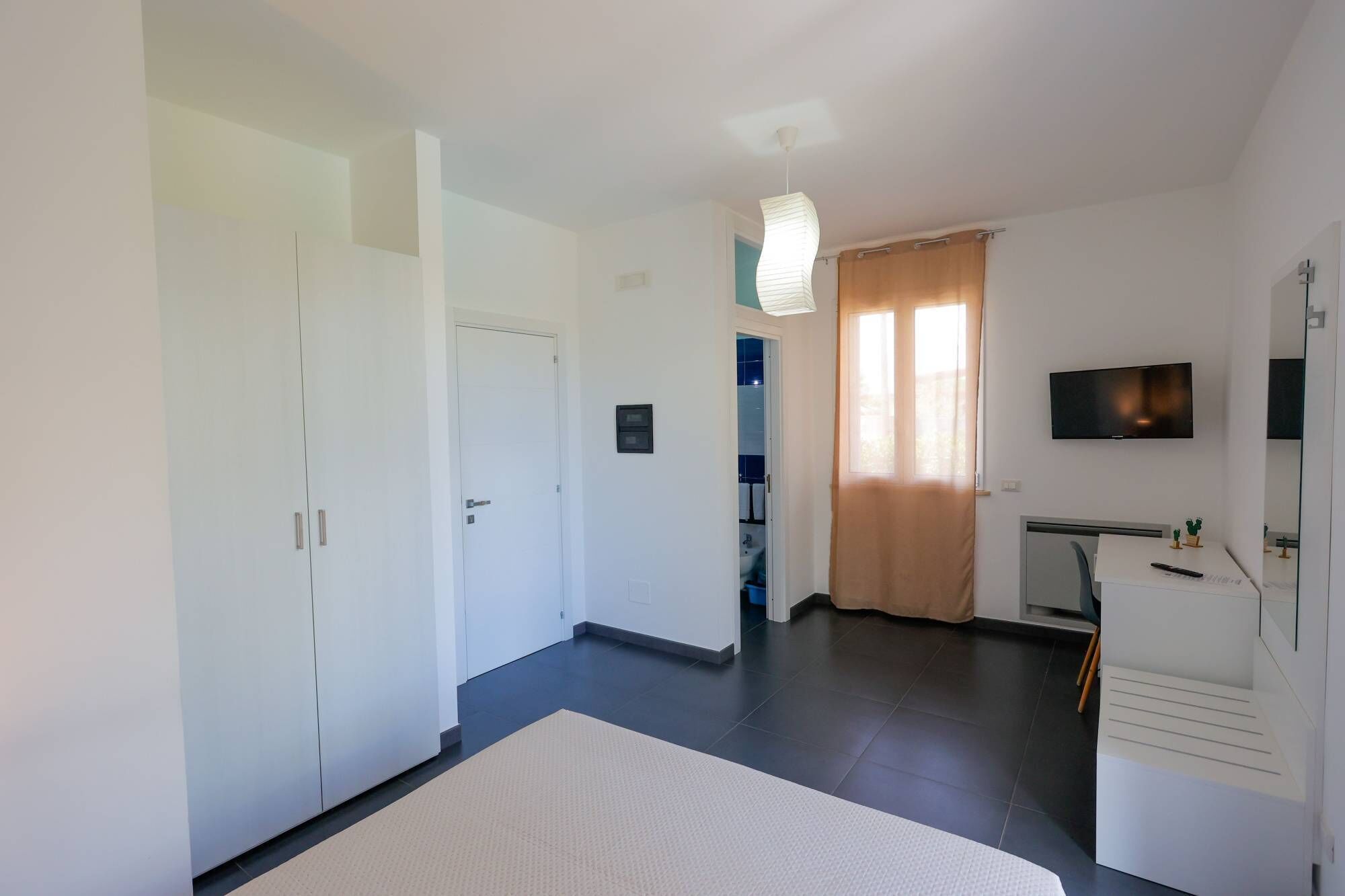 A modern hotel room with a flat-screen TV, a desk with a chair, a bed, a wardrobe, and a bathroom visible through an open door.