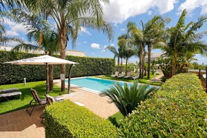 Una piscina rodeada de palmeras y un seto, con sillas de descanso y una pérgola.