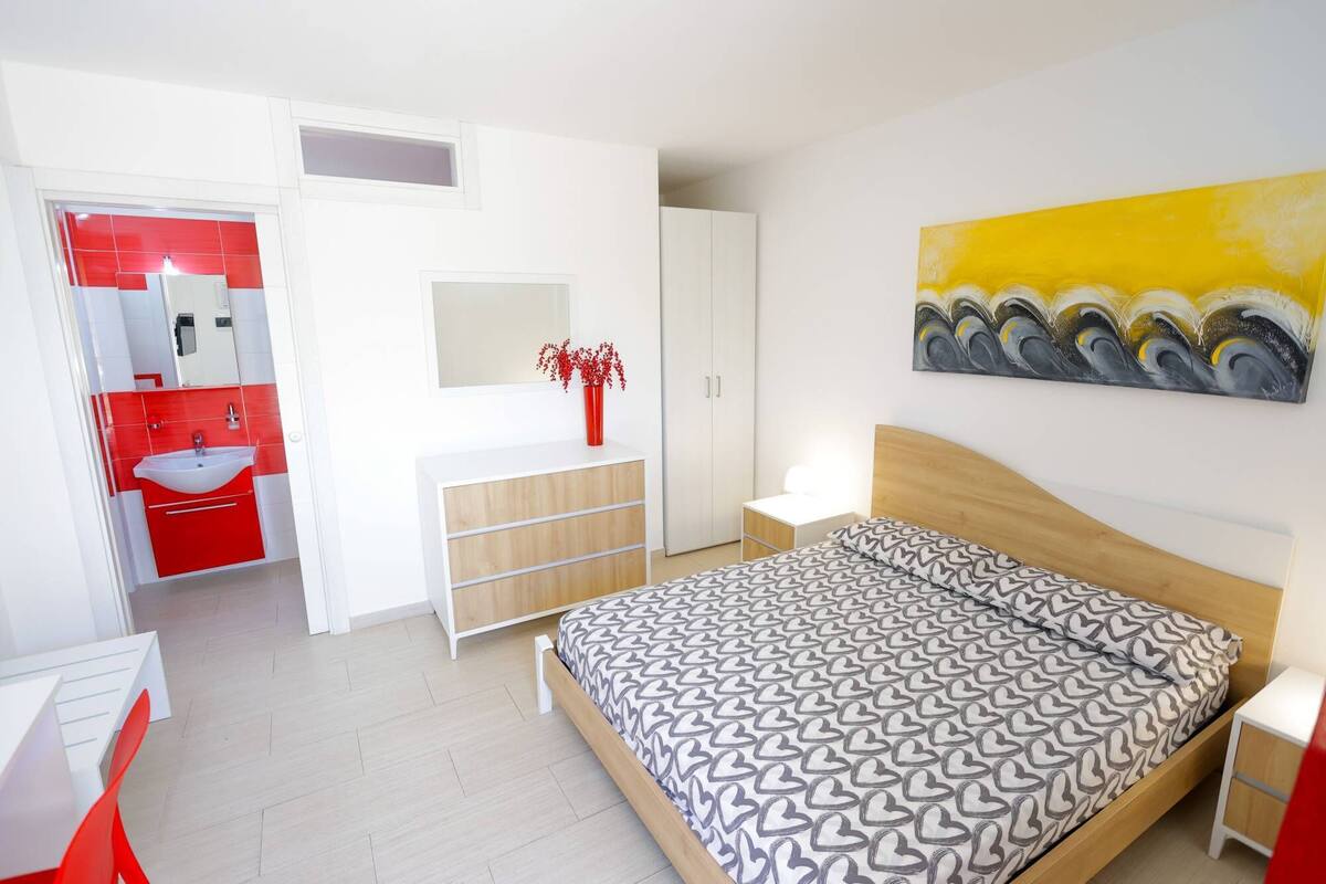 A bedroom with a bed, a bedside table, a nightstand, a wardrobe, a mirror, and a bathroom with a sink and a red cabinet.