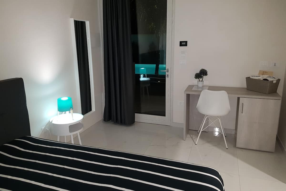 A hotel room with a bed, a desk, a chair, a small table with a lamp, a potted plant, and a cabinet.