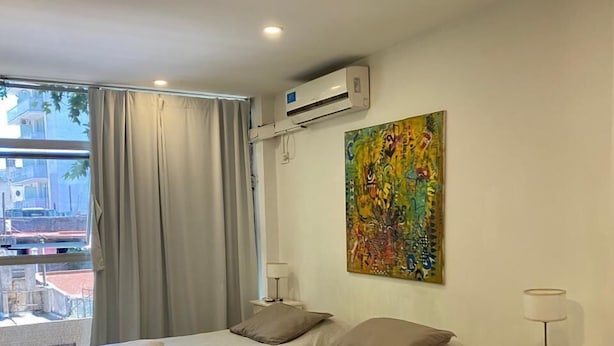 A hotel room with two beds, an air conditioner, a painting, a desk, and a view of the outside.