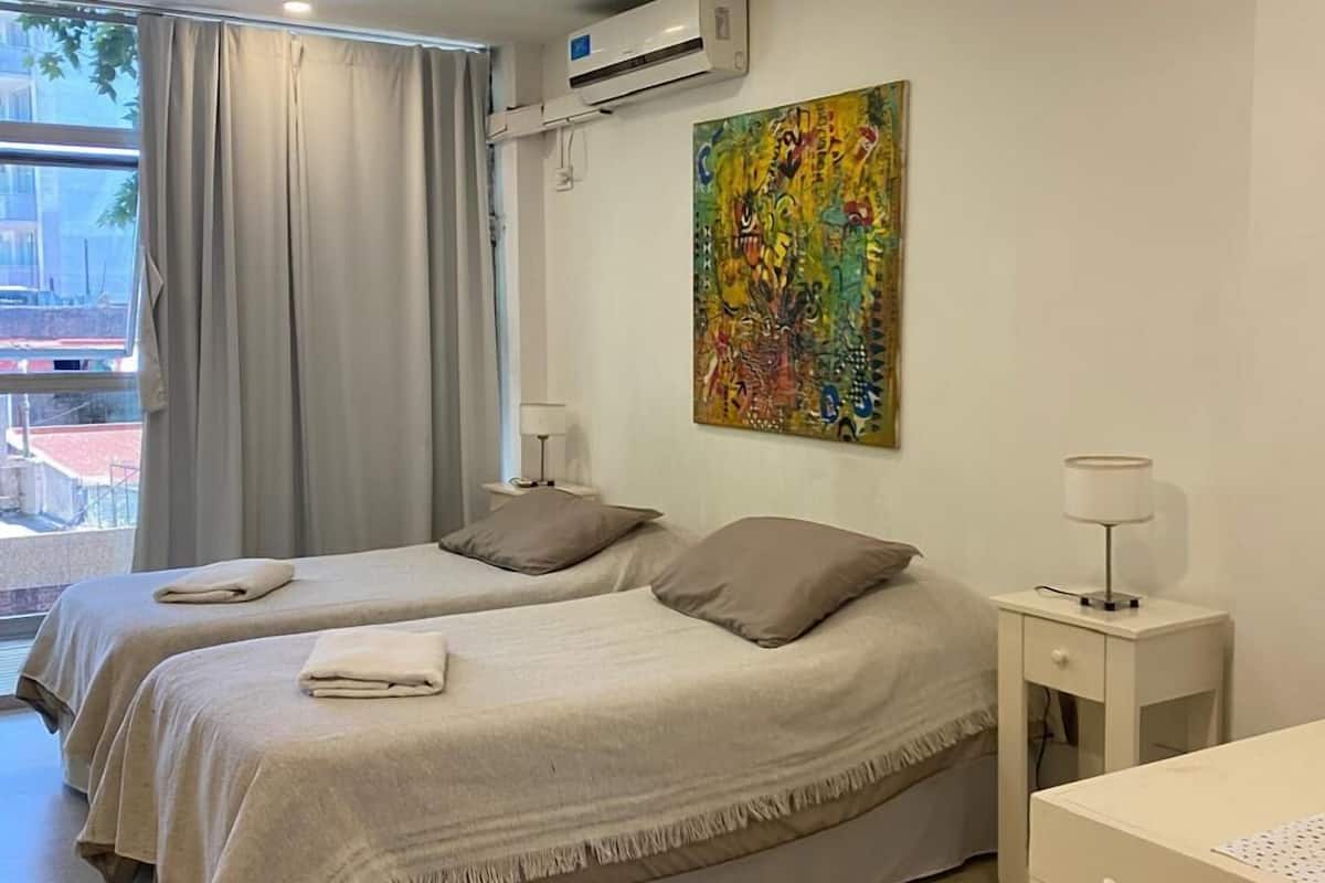A hotel room with two beds, a desk, a chair, an air conditioner, a window with curtains, and a colorful painting on the wall.