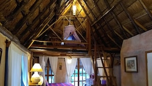 A rustic room with a thatched roof, a bed with a colorful blanket, a small table with a lamp, and a window with curtains. - Casitas Kinsol (Puerto Morelos)