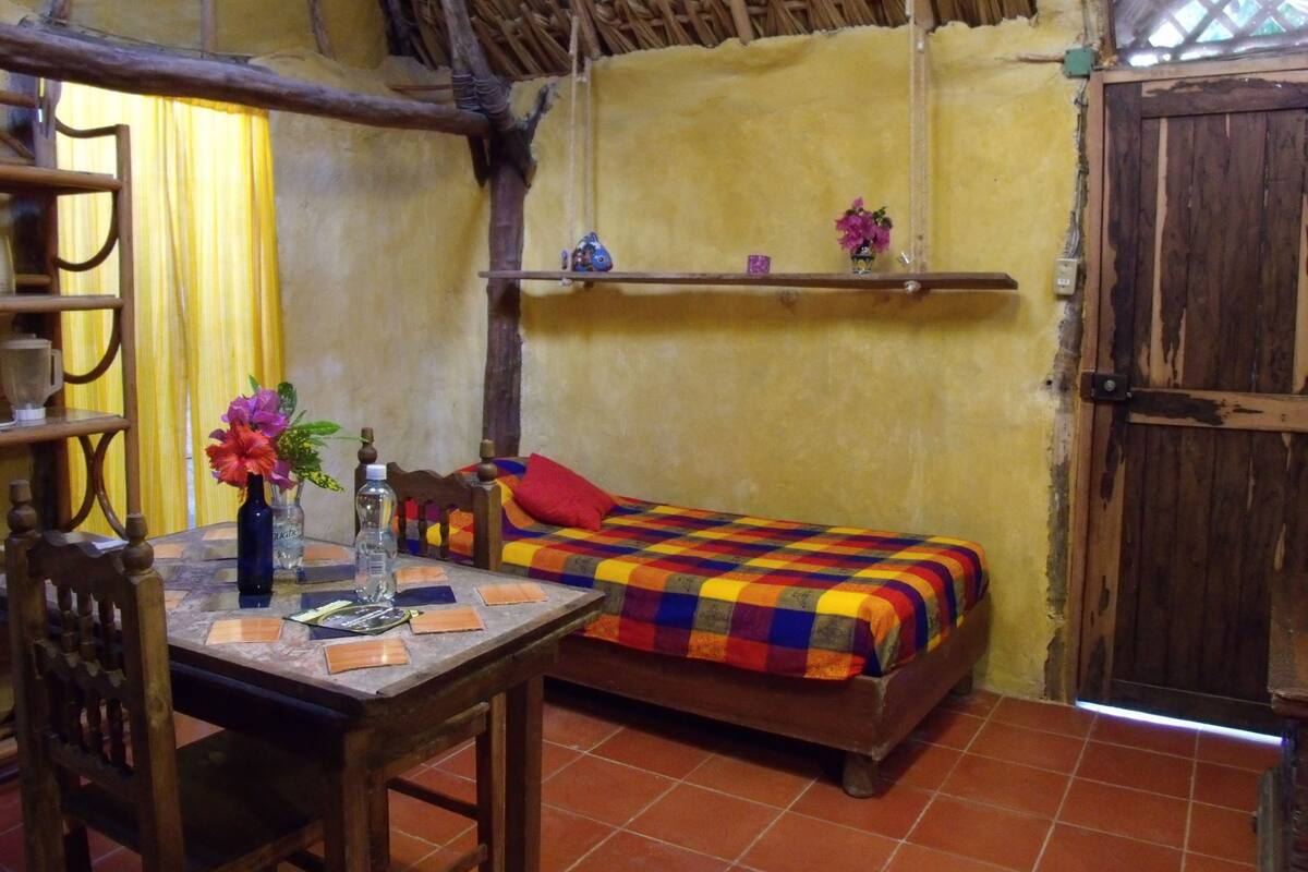 Una habitación rústica con una cama de madera, una mesita con un florero y una silla.