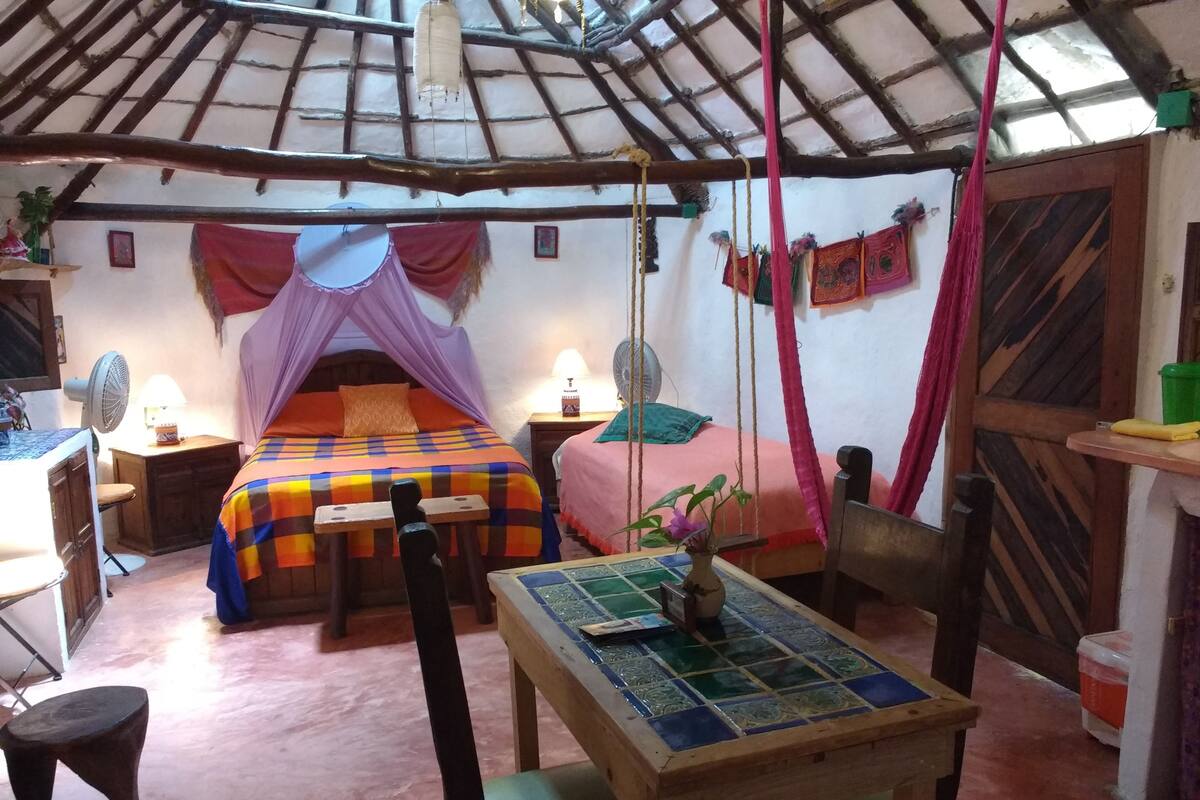 Una habitación con una cama bajo un techo de paja, una mesa con cubierta de azulejos y una silla.