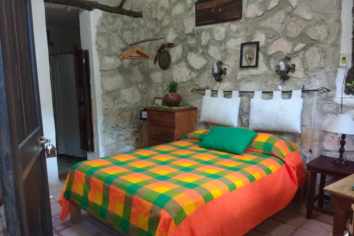 Un dormitorio con pared de piedra, una cama con colcha de cuadros coloridos, una mesita de noche de madera con una lámpara y un perchero.
