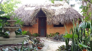 A rustic thatched-roof house with a small window and a door, surrounded by lush greenery and a stone fountain. - Casitas Kinsol (Puerto Morelos)