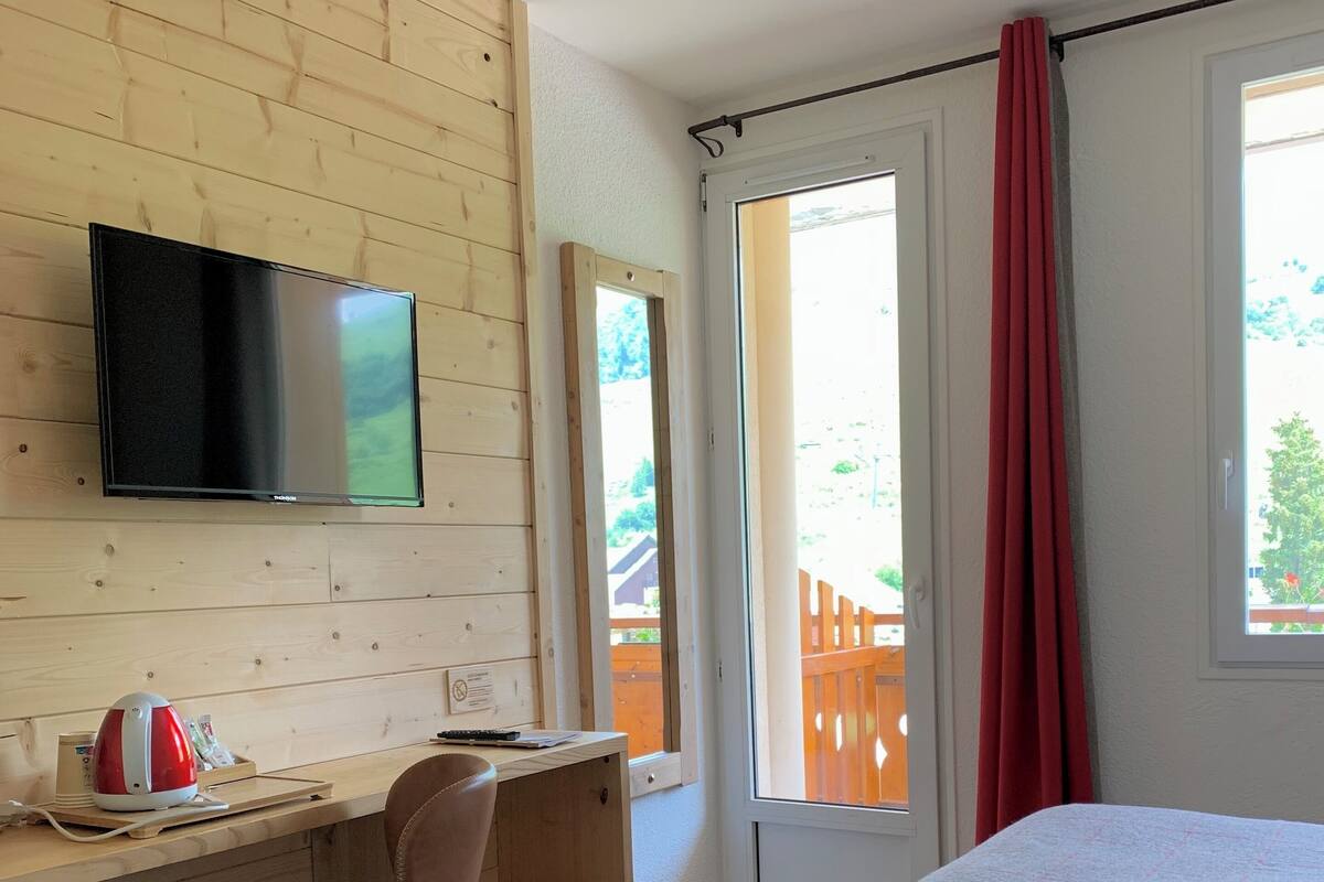 Een slaapkamer met een bed, een bureau met stoel, een televisie en een raam met uitzicht op een houten terras.