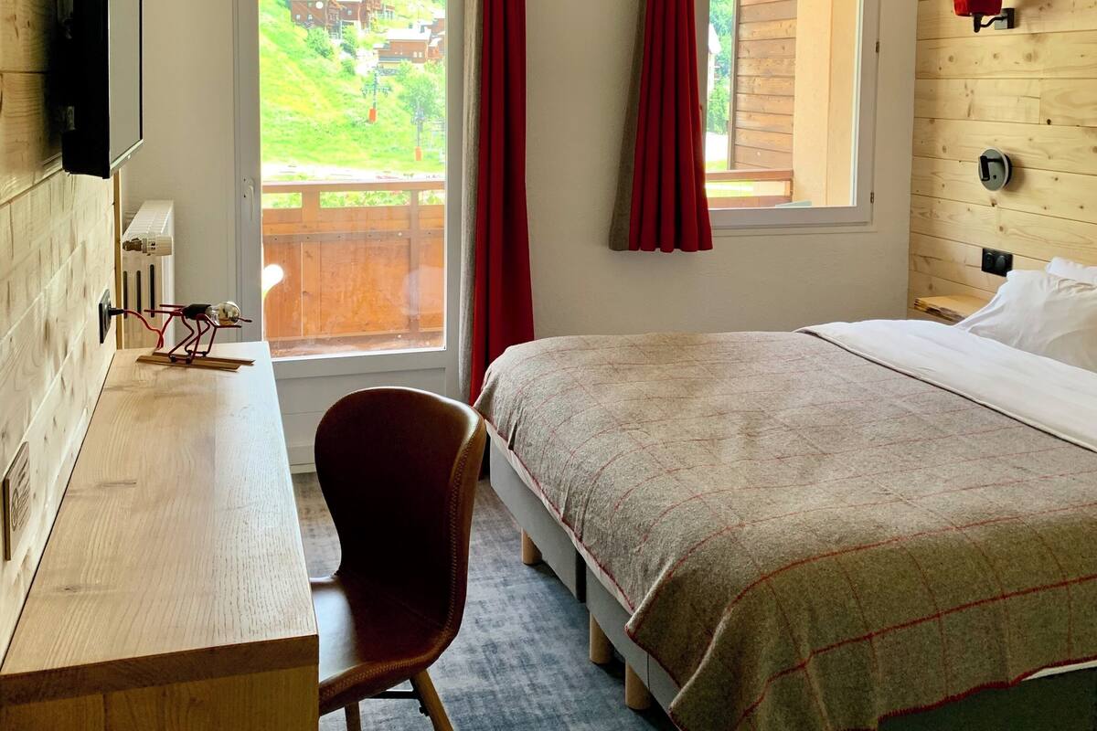 Een slaapkamer met een bed, een bureau, een stoel, een raam met gordijnen en uitzicht op een groen landschap.