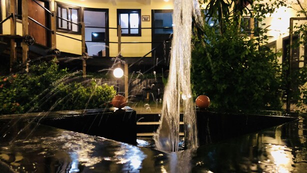 A fountain with water jets and two spherical ornaments, surrounded by greenery and a building with windows and a door. - Détente Hôtel (Abidjan)