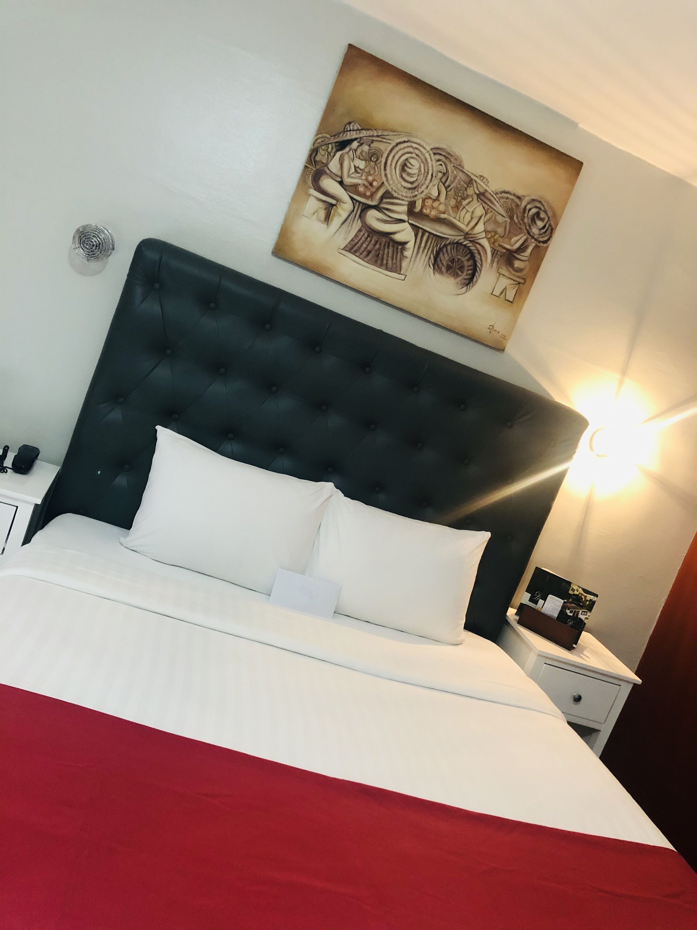 A hotel room with a large bed, a dark headboard, a bedside table with a lamp, and a wall-mounted artwork.