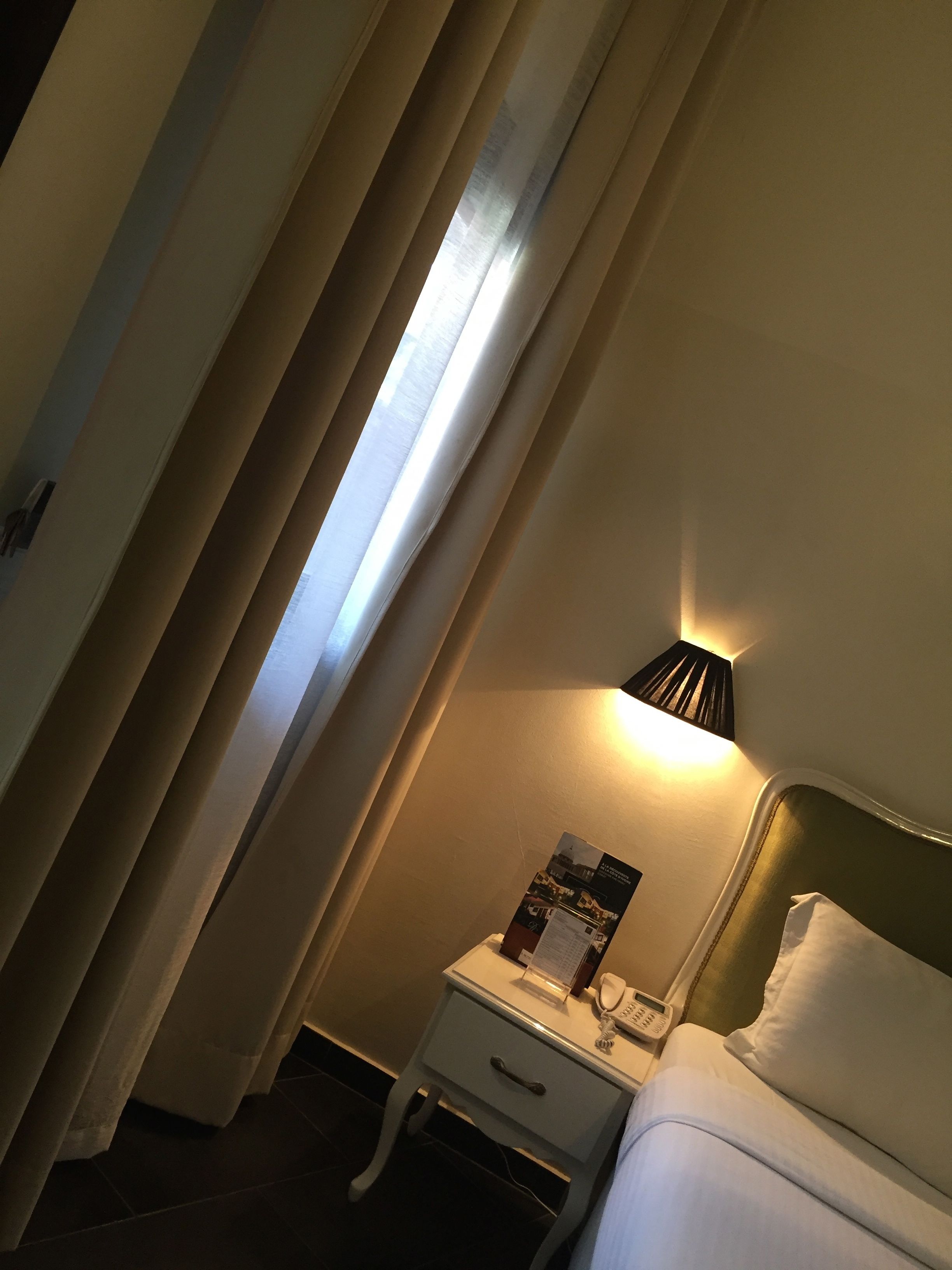A hotel room with a bed, a bedside table with a lamp, a telephone, and a poster on the wall.