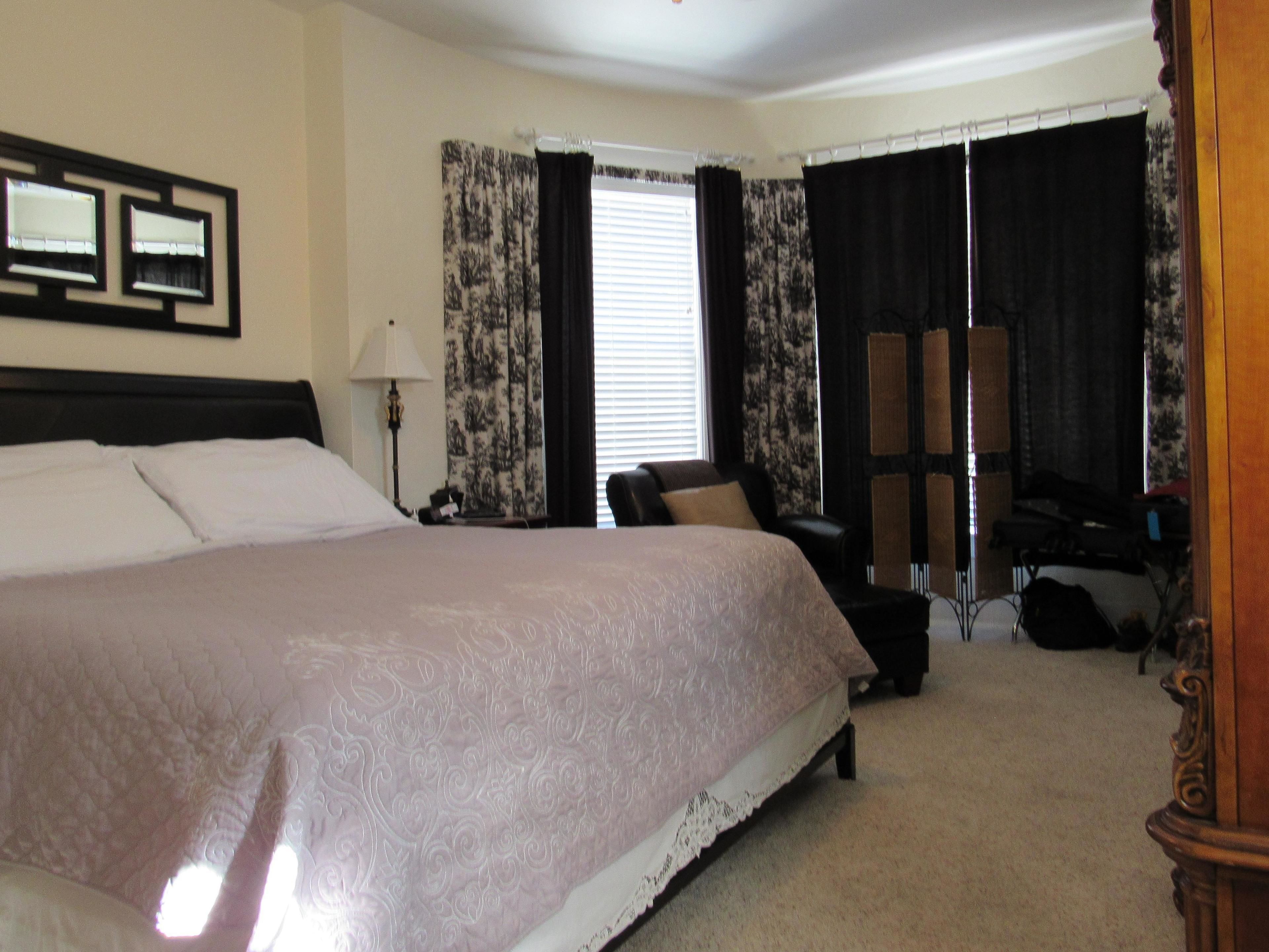 A spacious bedroom with a large bed, a desk, and a chair. There is a window with curtains, a lamp, and a mirror on the wall.