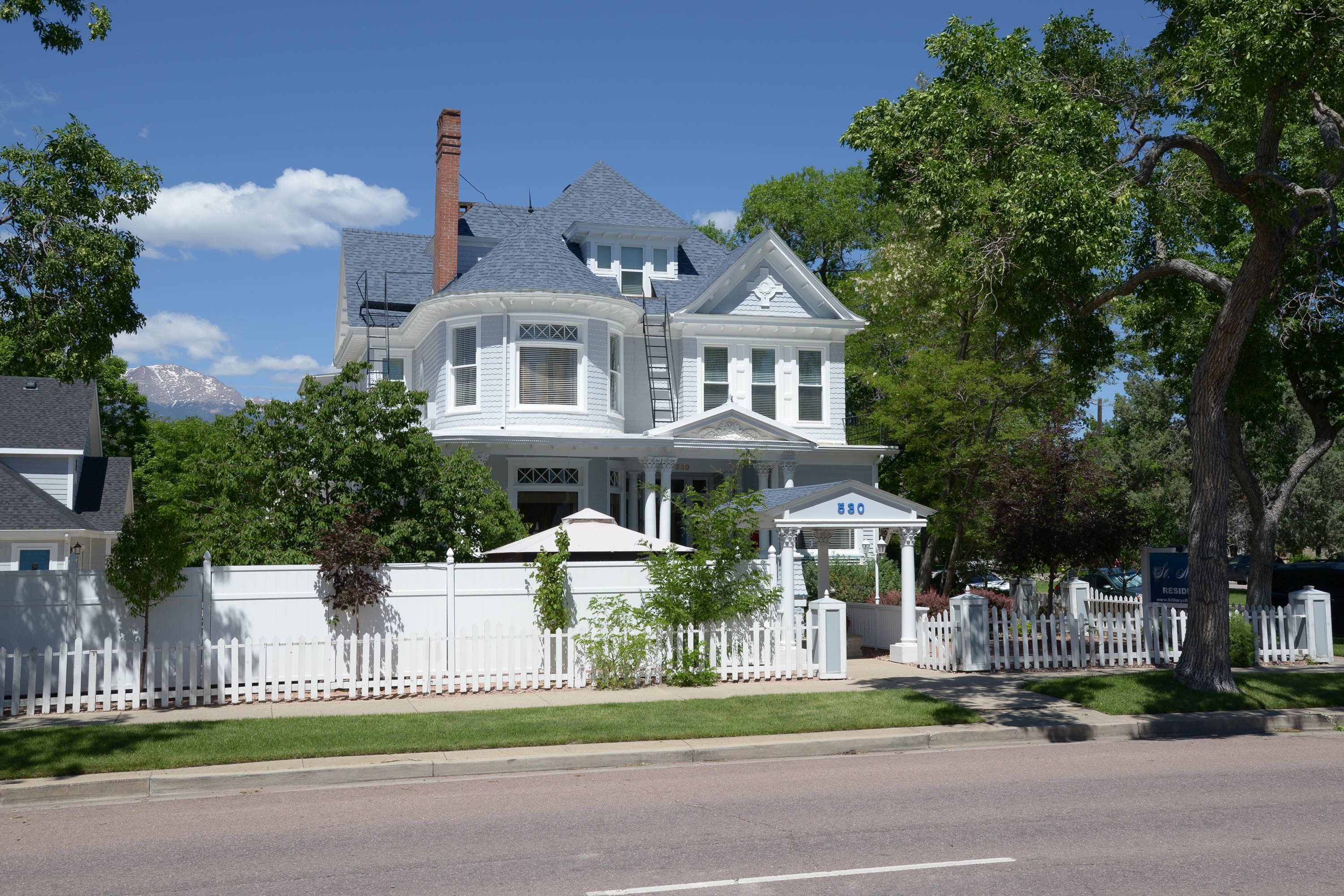 A large, two-story house with a prominent chimney, a white picket fence, and a well-maintained lawn.